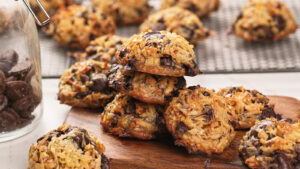 A stack of magic cookies on a wooden board, with more cookies cooling on a wire rack and a glass jar of chocolate pieces nearby.