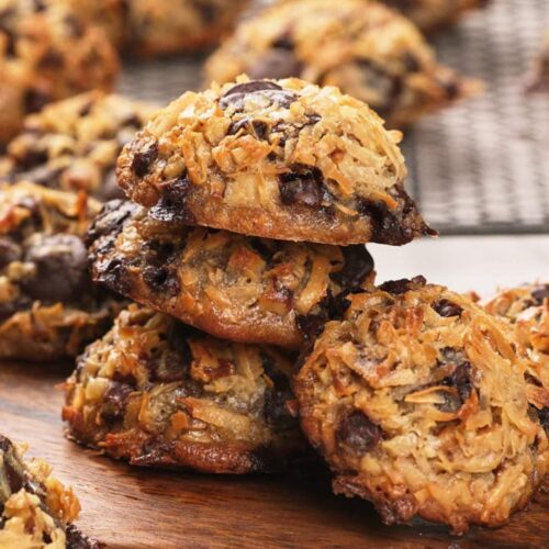 A stack of magic cookies on a wooden board, with more cookies cooling on a wire rack in the background.