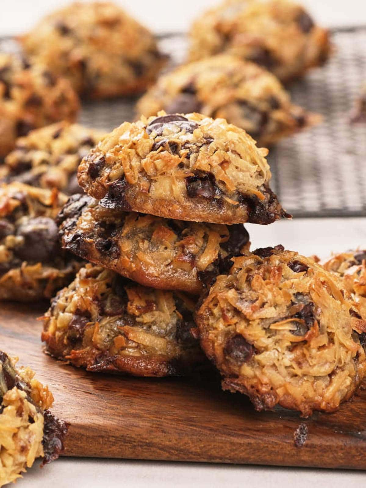 A stack of magic cookies on a wooden board, with more cookies cooling on a wire rack in the background.
