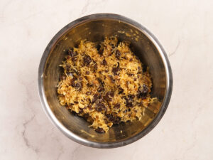 A metal bowl containing a mixture of shredded coconut, raisins, and other baking ingredients on a light countertop.
