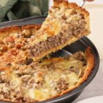 A hand lifts a slice of savory ground meat and cheese pie from a round baking pan, showing a golden crust and melted cheese topping.