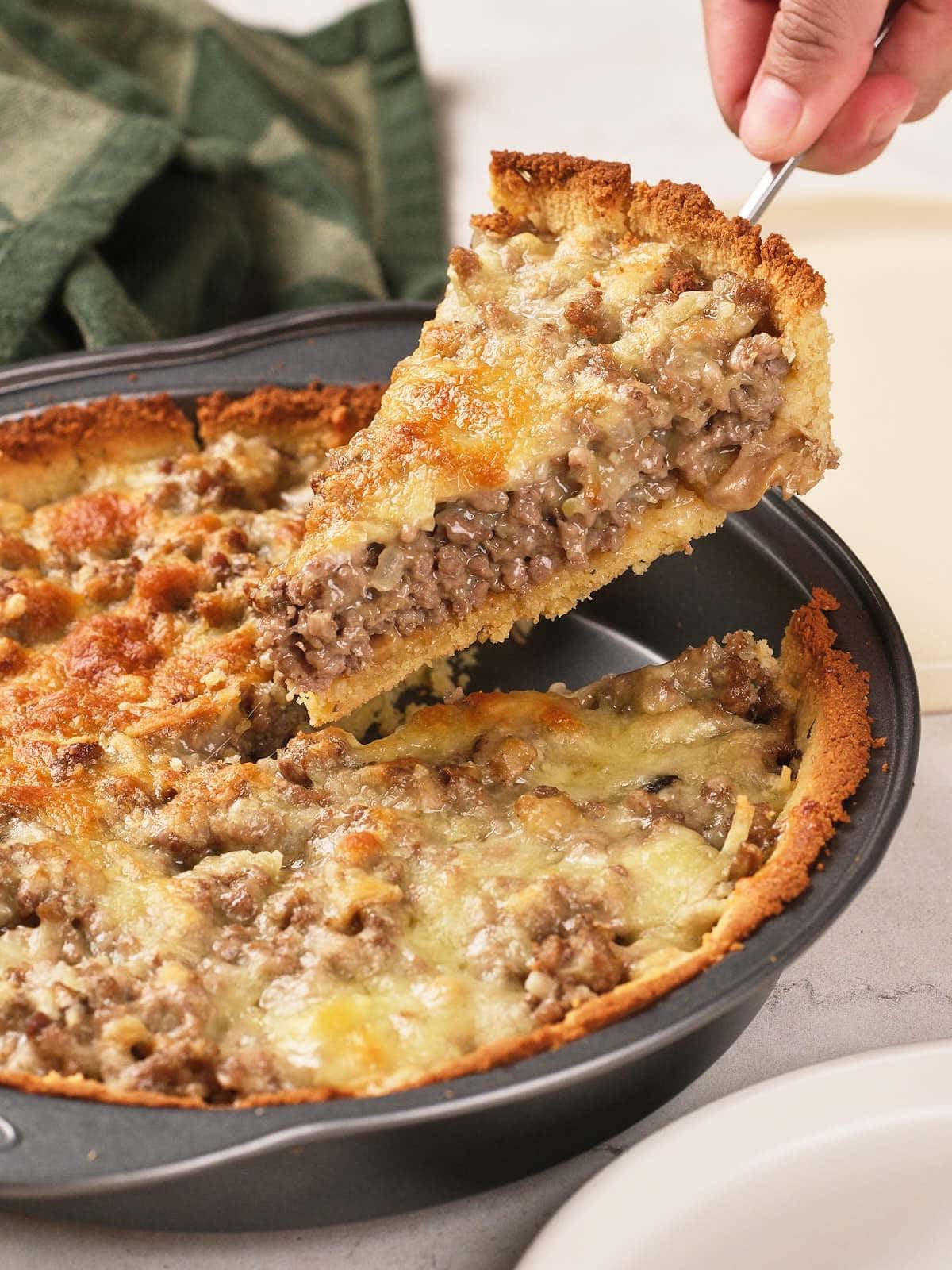 A hand lifts a slice of savory ground meat and cheese pie from a round baking pan, showing a golden crust and melted cheese topping.