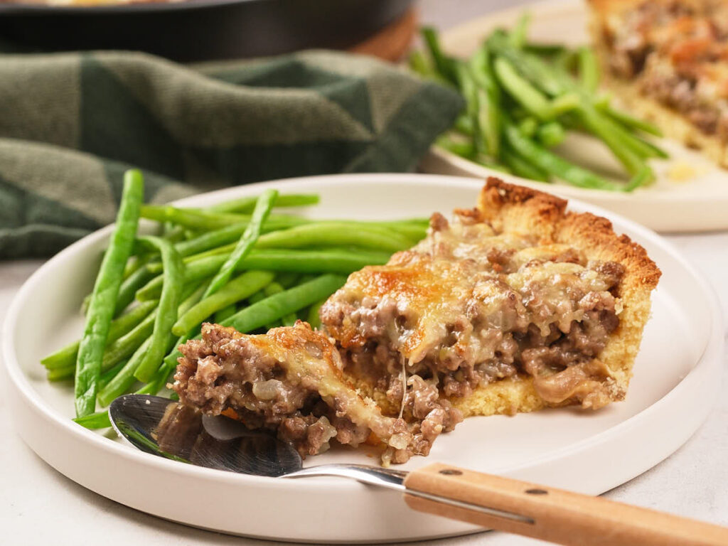 A slice of savory meat pie with melted cheese served alongside cooked green beans on a white plate with a spoon and fork.