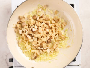 Chopped mushrooms and diced onions cooking in a light-colored frying pan on a stove.