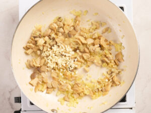 Chopped mushrooms, diced onions, and minced garlic cooking in a white skillet on a stovetop.