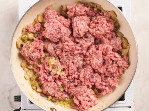 Raw ground meat and chopped onions are cooking in a skillet on a stovetop.