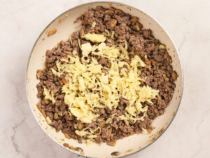 Ground beef and sautéed mushrooms cooked in a skillet, topped with a pile of shredded cheese, on a light-colored countertop.