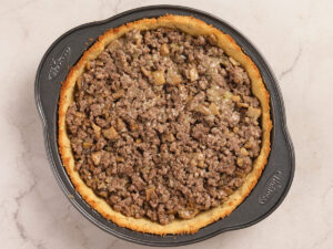 A pie pan filled with a baked crust and topped with a mixture of cooked ground beef and diced mushrooms.