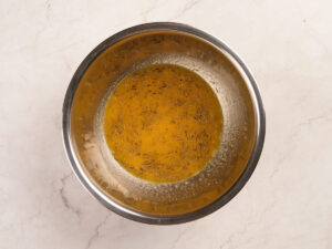 A metal bowl containing a yellow liquid with visible herb particles on a light-colored countertop.