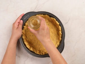 Hands pressing dough into a round baking pan using the bottom of a glass jar on a light-colored countertop.