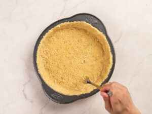 A hand uses a fork to poke holes in an unbaked pie crust in a round baking pan on a light countertop.
