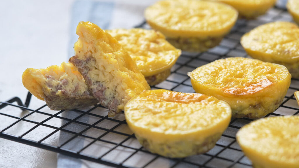Mini egg muffins with visible meat pieces are cooling on a black wire rack; one muffin is split open, showing the inside texture.