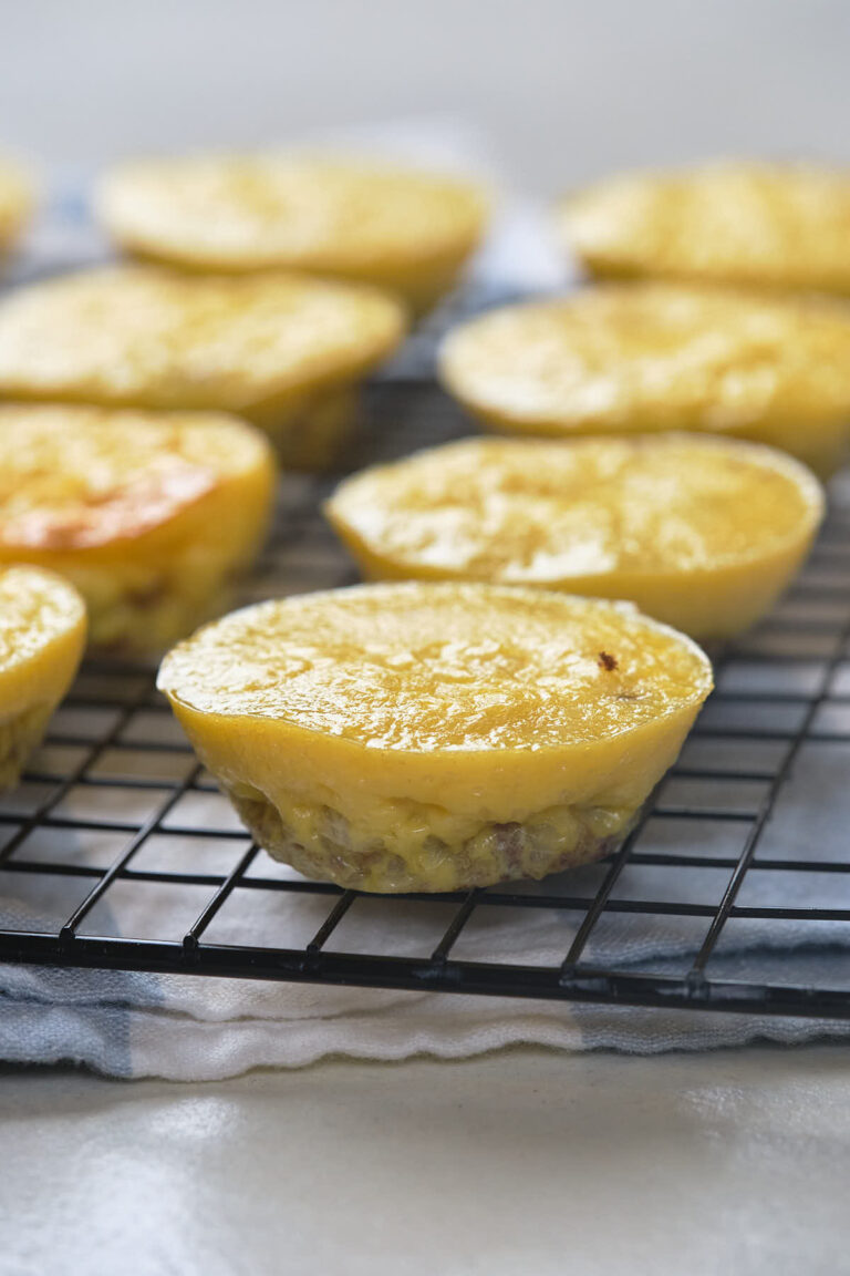 Mini egg muffins cooling on a black wire rack, with a light cloth underneath and more egg muffins in the background.