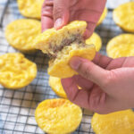A person holds a half-eaten yellow pastry with meat filling above a cooling rack with several whole pastries.