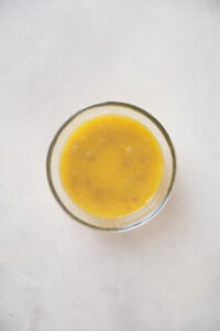 A small glass bowl filled with a yellow liquid mixture containing visible small brown specks, placed on a light-colored surface.