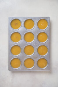 A muffin tin with twelve cups filled with a yellow liquid mixture, placed on a light-colored surface.