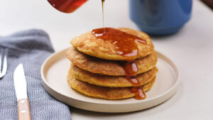 A stack of pancakes on a plate with syrup being poured on top, next to a knife and a blue cloth napkin.