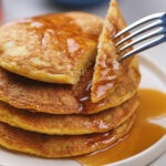 A fork lifts a bite from a stack of pancakes covered in syrup on a white plate.
