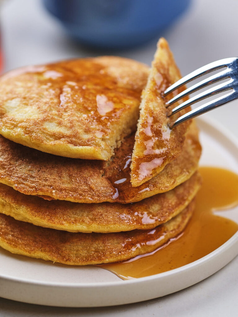 A fork lifts a bite from a stack of pancakes covered in syrup on a white plate.