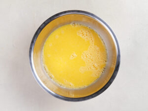 A stainless steel bowl containing a yellow liquid mixture with bubbles on the surface, placed on a light-colored countertop.