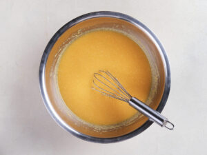 A metal mixing bowl filled with yellow batter and a metal whisk resting inside, on a light-colored surface.