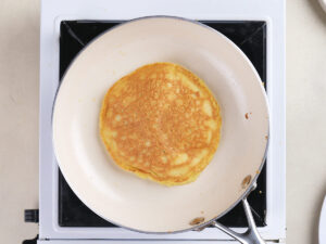 A golden-brown pancake cooking in a white frying pan on a stovetop.