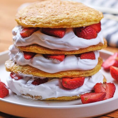 Three layers of shortcake with whipped cream and sliced strawberries, stacked on a white plate with extra strawberry pieces on the side.