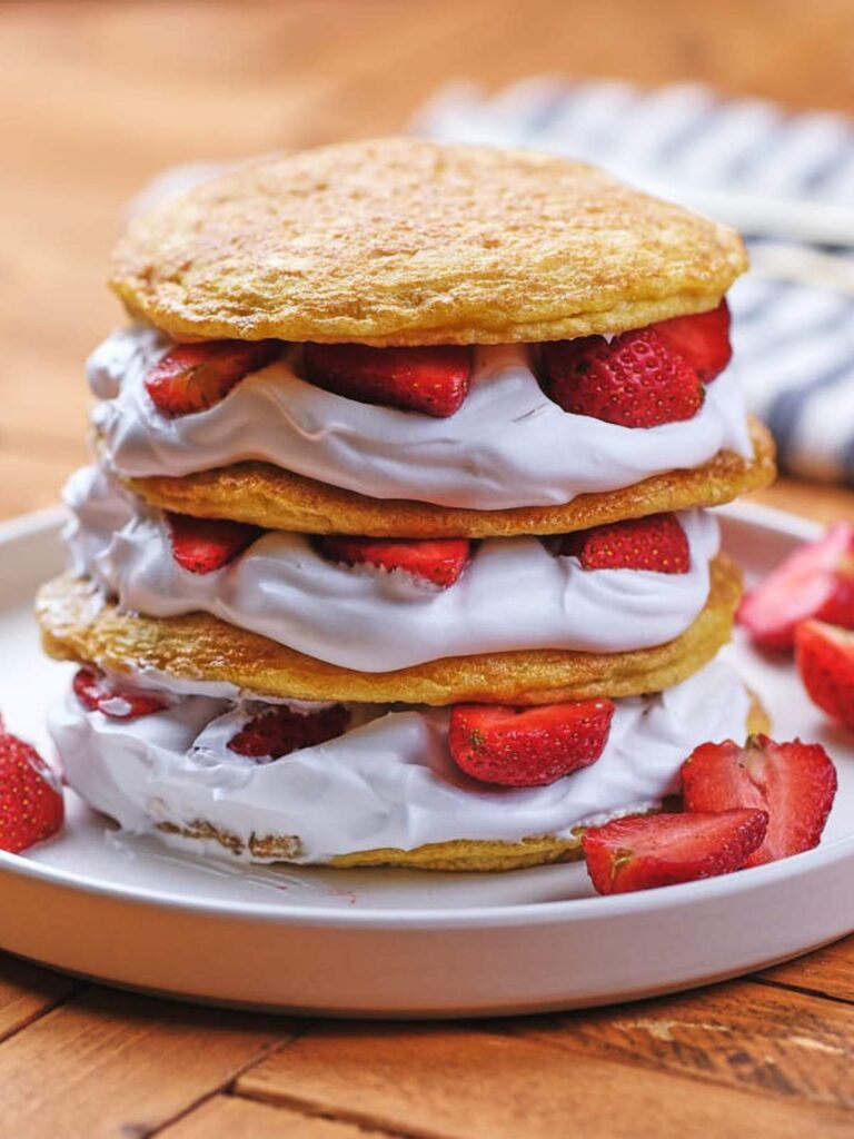 Three layers of shortcake with whipped cream and sliced strawberries, stacked on a white plate with extra strawberry pieces on the side.