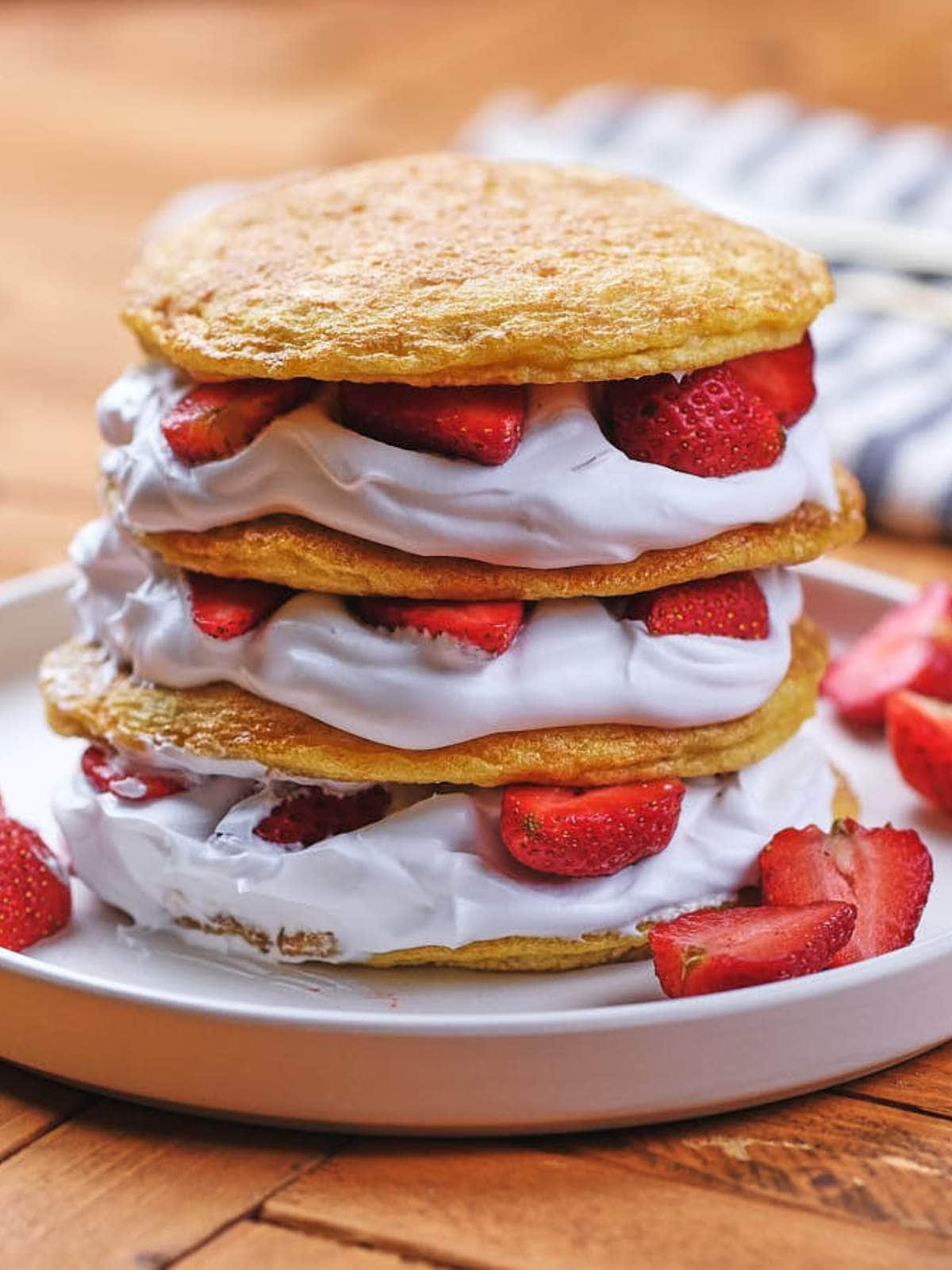Three layers of shortcake with whipped cream and sliced strawberries, stacked on a white plate with extra strawberry pieces on the side.