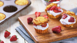 Two shortcakes filled with whipped cream and sliced strawberries are on a wooden board, with fresh strawberries and a muffin tin in the background.