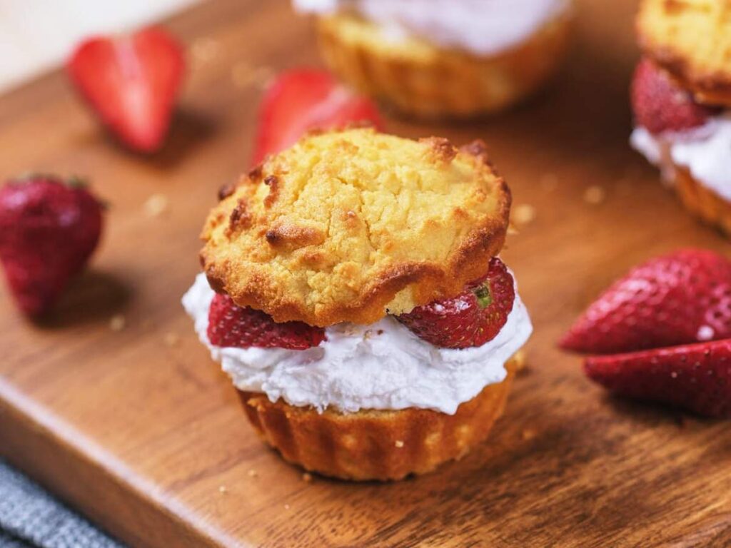 A strawberry shortcake with whipped cream and fresh strawberries sits on a wooden board, with additional strawberries in the background.