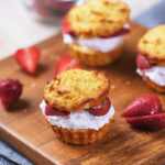 Three shortcake biscuits filled with whipped cream and sliced strawberries on a wooden board, with additional strawberries nearby.