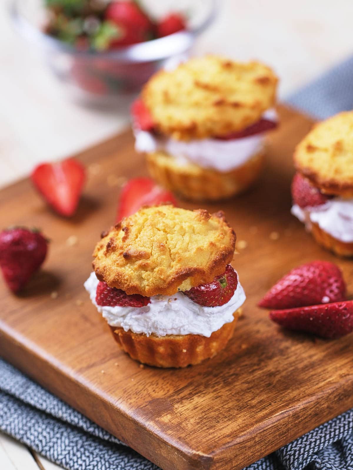 Three shortcake biscuits filled with whipped cream and sliced strawberries on a wooden board, with additional strawberries nearby.
