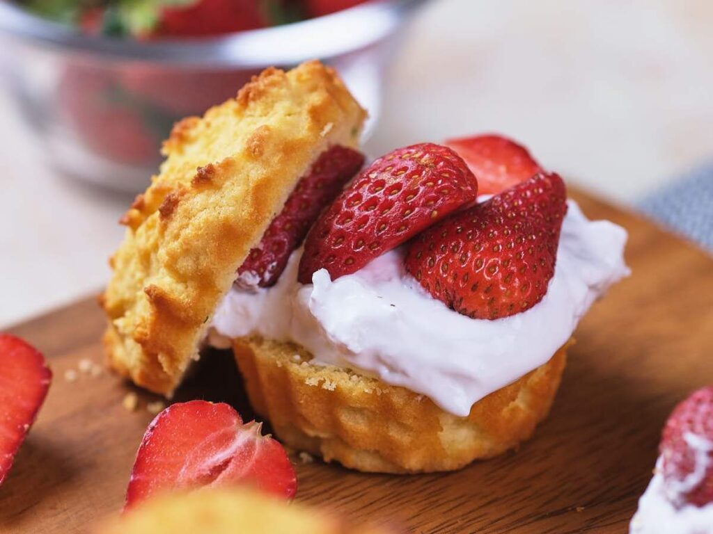 A biscuit split in half and filled with whipped cream and sliced strawberries, placed on a wooden board with more strawberries in the background.