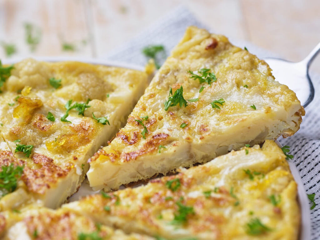 A close-up of a sliced cauliflower frittata garnished with chopped parsley on a white plate.