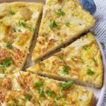 A sliced vegetable frittata garnished with chopped herbs is arranged on a white plate with a serving spoon nearby.