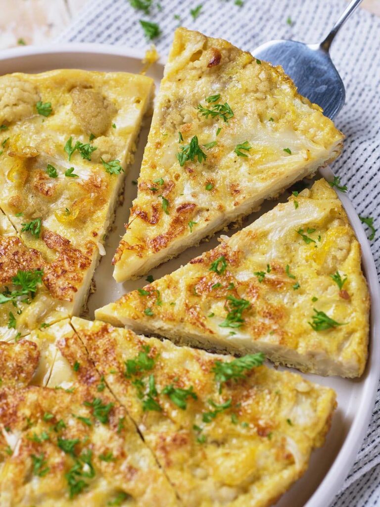 A sliced vegetable frittata garnished with chopped herbs is arranged on a white plate with a serving spoon nearby.