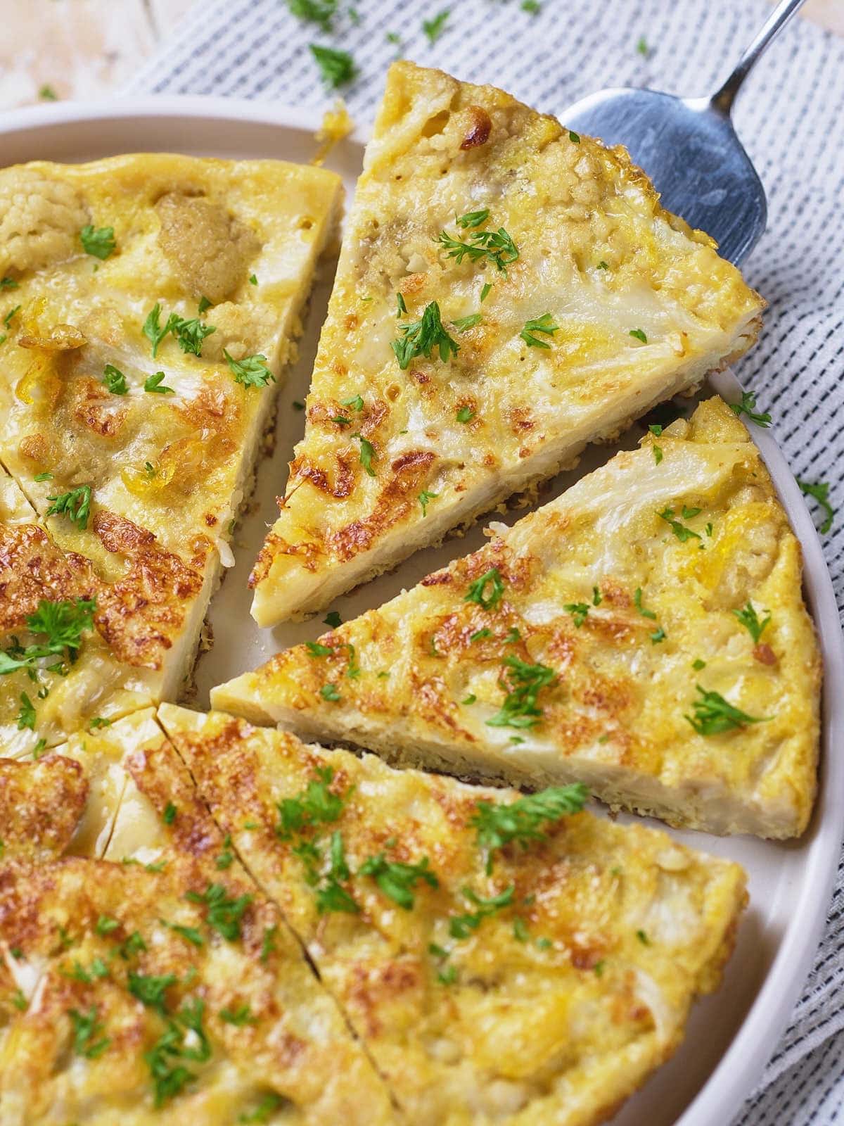A sliced vegetable frittata garnished with chopped herbs is arranged on a white plate with a serving spoon nearby.
