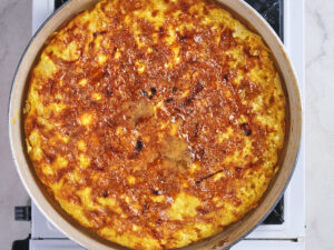 A large, golden-brown Spanish omelette is cooking in a metal skillet on a stovetop, viewed from above.