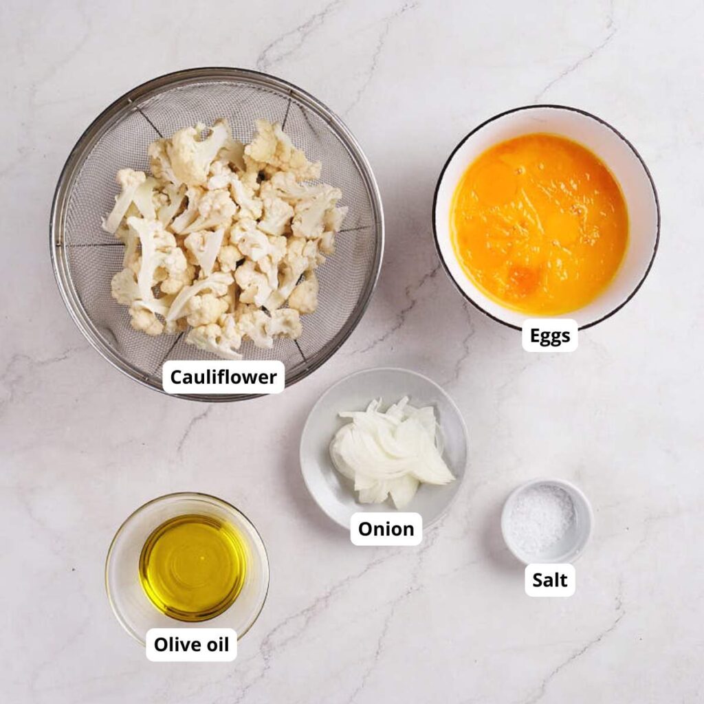 Five labeled bowls containing cauliflower florets, beaten eggs, sliced onion, olive oil, and salt are arranged on a light-colored countertop.