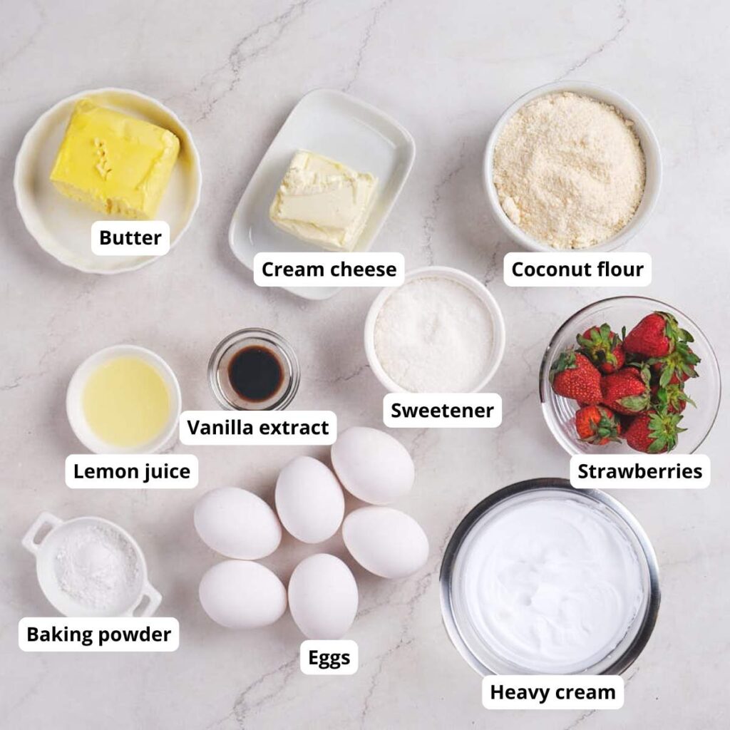 Various baking ingredients including butter, cream cheese, coconut flour, sweetener, strawberries, lemon juice, vanilla extract, eggs, baking powder, and heavy cream on a countertop.