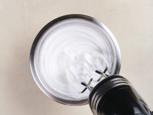 A hand mixer whipping egg whites into stiff peaks in a stainless steel bowl on a light-colored surface.