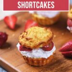 A mini strawberry shortcake with whipped cream and fresh strawberry slices is displayed on a wooden board with more shortcakes and strawberries in the background. Text reads: "Low Carb Mini Strawberry Shortcakes.