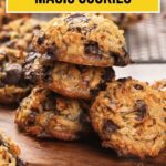 A stack of sugar-free magic cookies with coconut and chocolate chips on a wooden board, with more cookies and a cooling rack in the background.