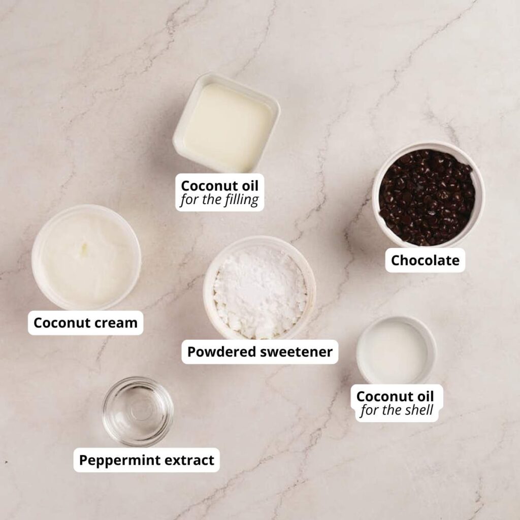 Six labeled ingredients on a marble surface: coconut cream, peppermint extract, powdered sweetener, coconut oil for filling, chocolate, and coconut oil for the shell.