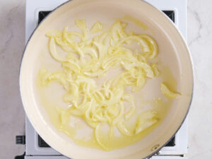 Sliced onions cooking in oil in a beige skillet on a stovetop.