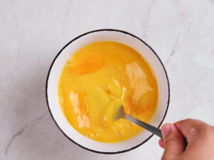 A hand uses a fork to whisk eggs in a white bowl on a light-colored countertop.