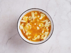 A bowl of cauliflower soup with visible florets and a yellowish broth, placed on a light marble surface.