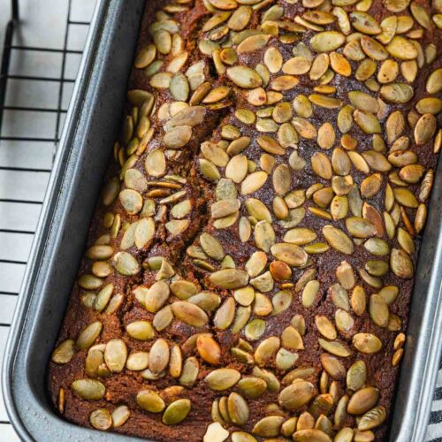 Loaf of quick bread topped with pumpkin seeds in a metal baking pan on a wire rack.