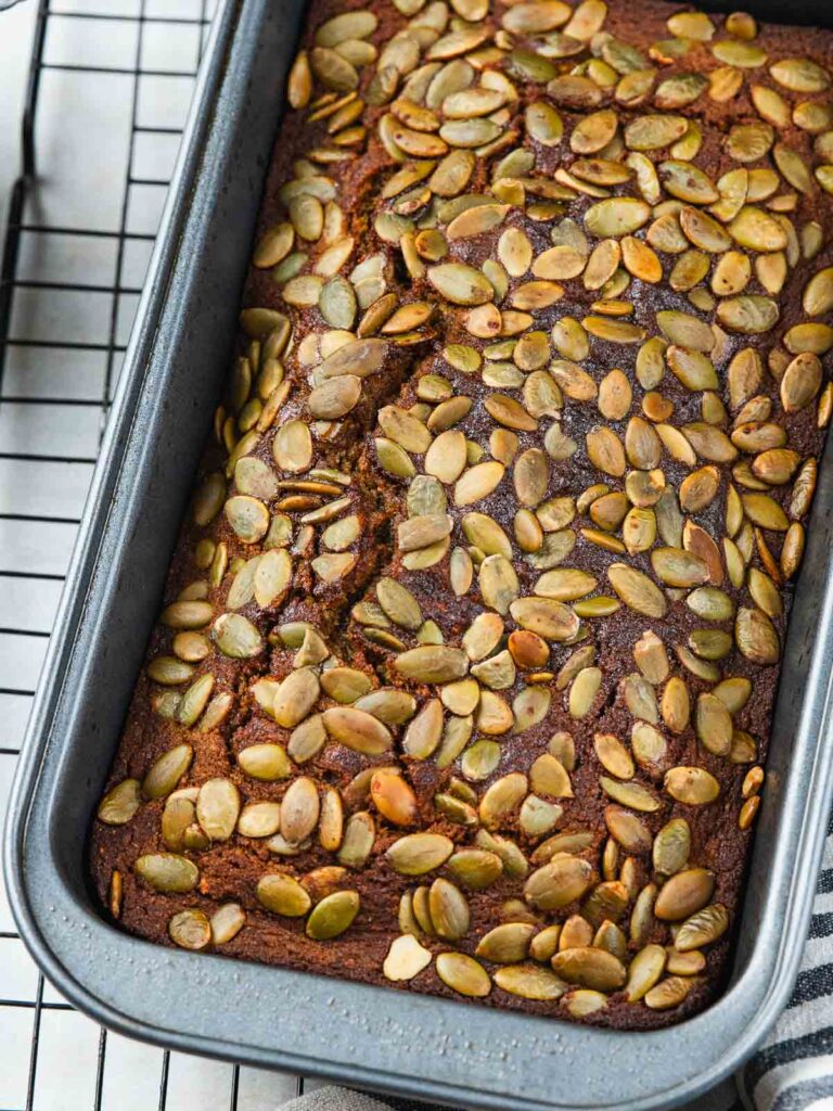 Loaf of quick bread topped with pumpkin seeds in a metal baking pan on a wire rack.